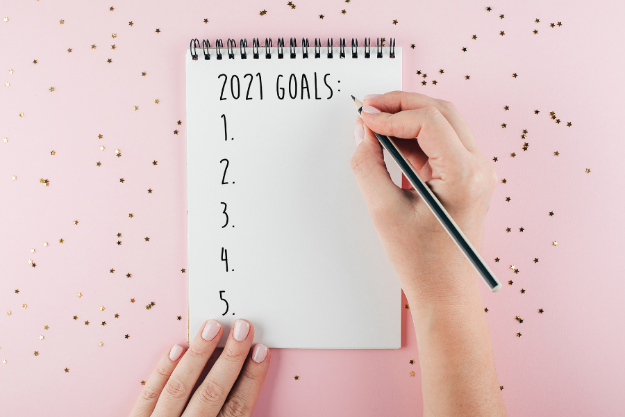 image of notepad sitting on pink background with glitter. Notepad says 2021 goals and hands are in the frame writing with a pen.